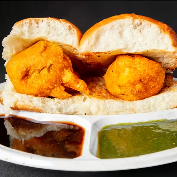 Two vada pavs, fried potato fritters in bread buns, served with green and brown chutneys.
