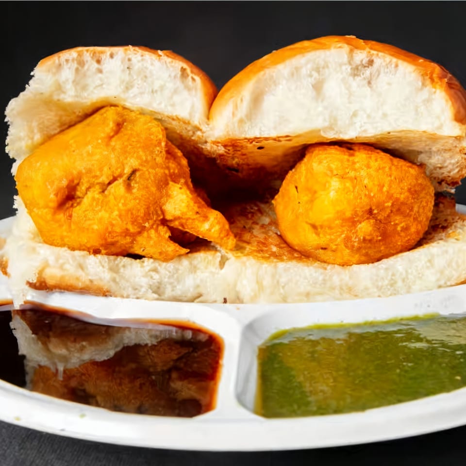 Two vada pavs, fried potato fritters in bread buns, served with green and brown chutneys.