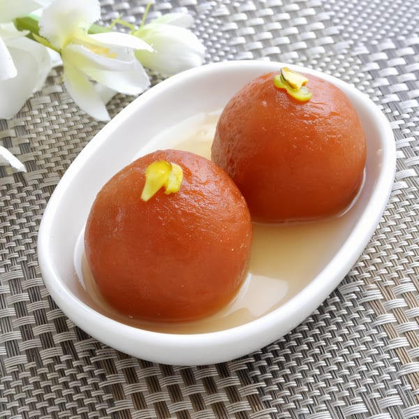 Two gulab jamun, garnished with pistachios, in a white dish with syrup on a woven mat.