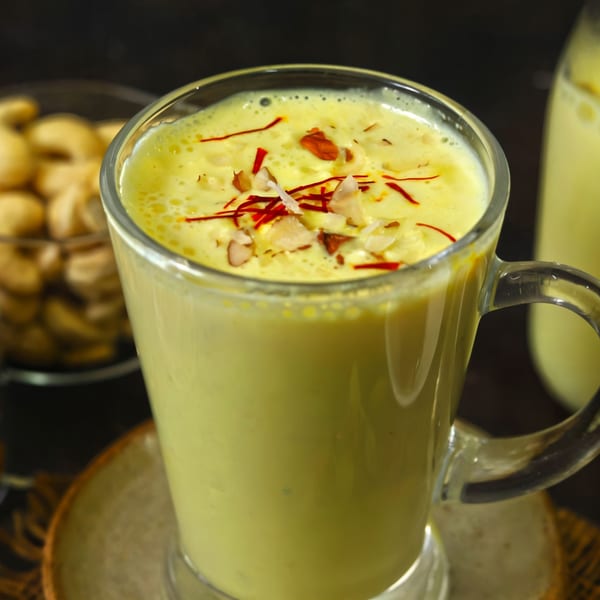 A glass of yellow Indian lassi, topped with saffron threads and chopped nuts, with cashews in the background.