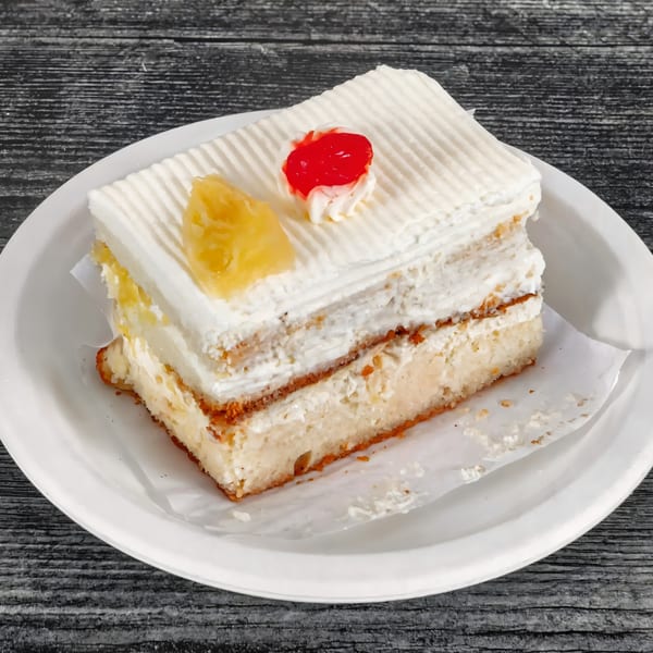 A slice of white cake with cream frosting, topped with pineapple and a cherry, on a white plate.